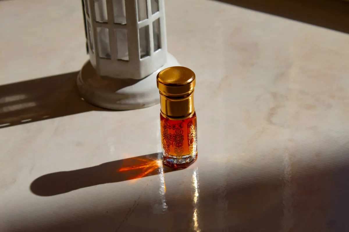 A gold-capped attar bottle on marble, throwing an amber light caustic beside a lantern.