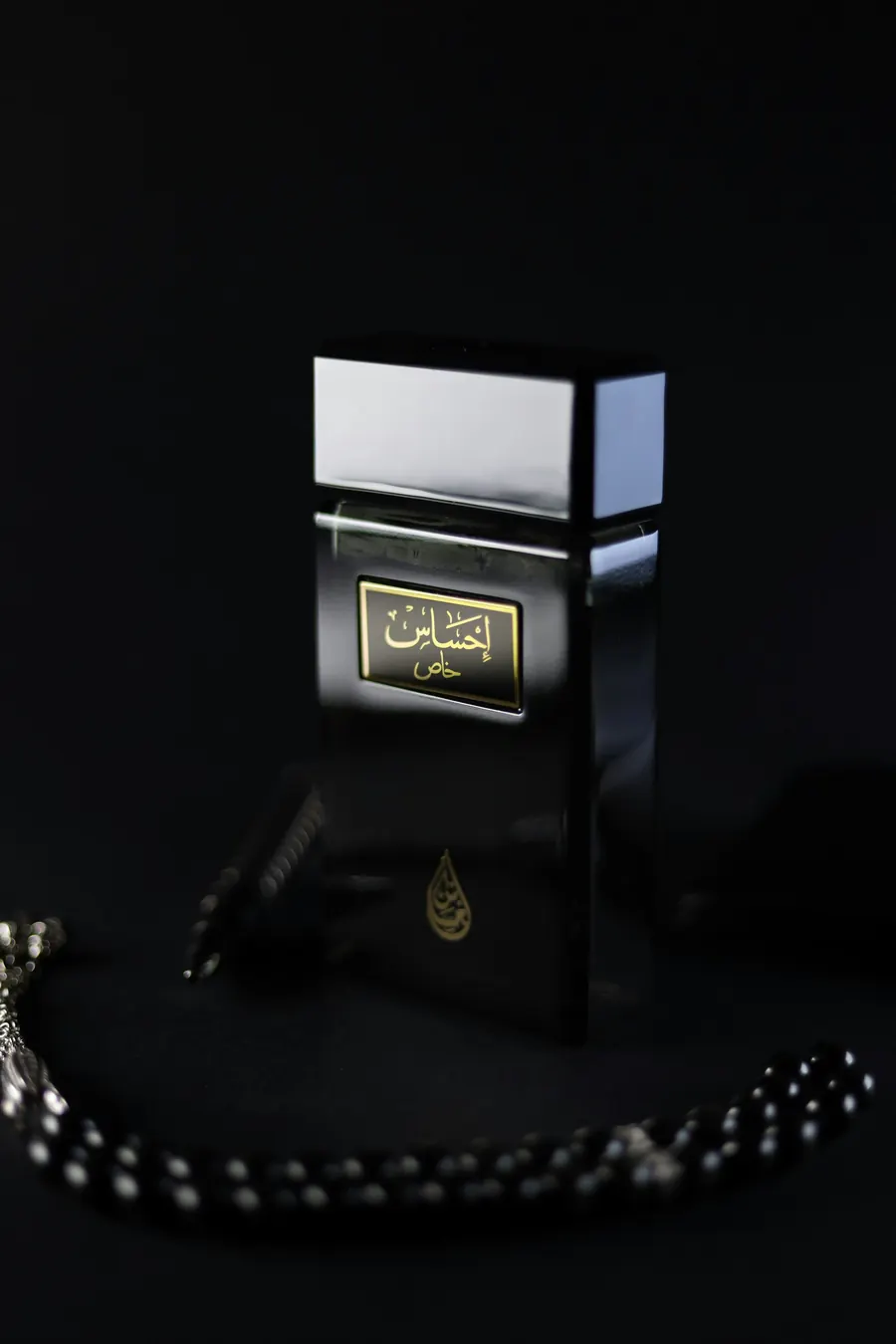 A black perfume bottle with a gold Arabic calligraphy label, resting beside prayer beads.